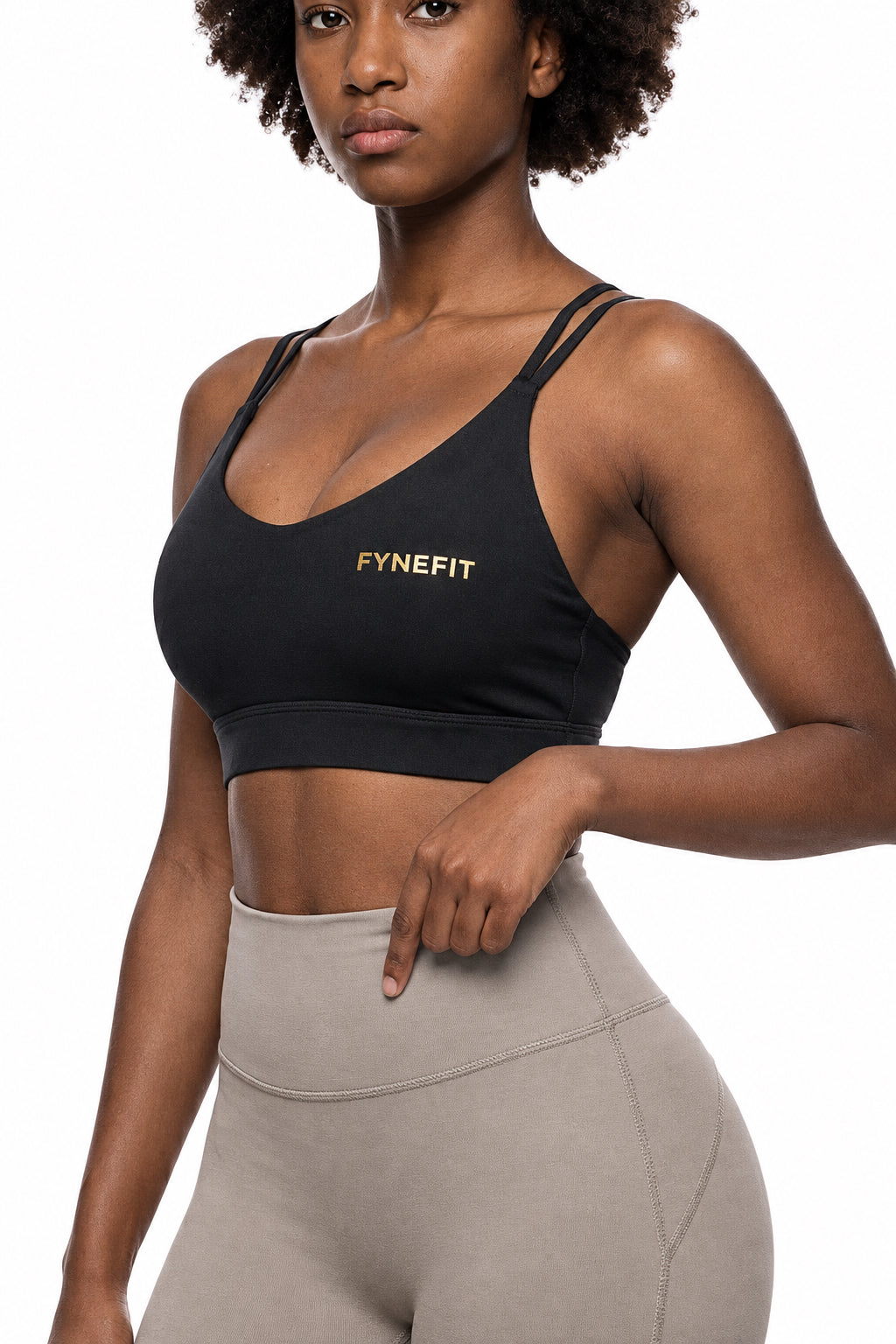 A woman modeling a black athletic sports bra with gold 'FYNEFIT' branding on the chest.